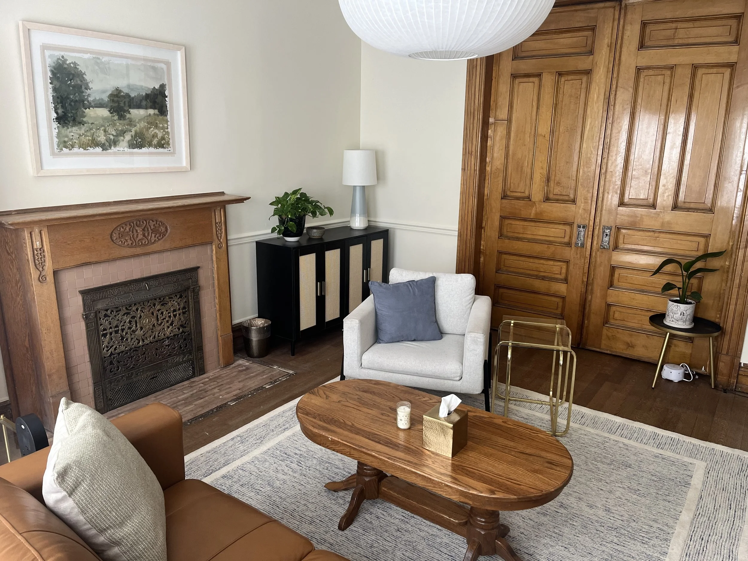 A cozy living room featuring a wooden fireplace with a decorative grate, a black cabinet with a potted plant and a white lamp, a white armchair with a blue pillow, a wooden coffee table, and a built-in wooden door closet. There's also a large paper lantern ceiling light and framed artwork on the wall.