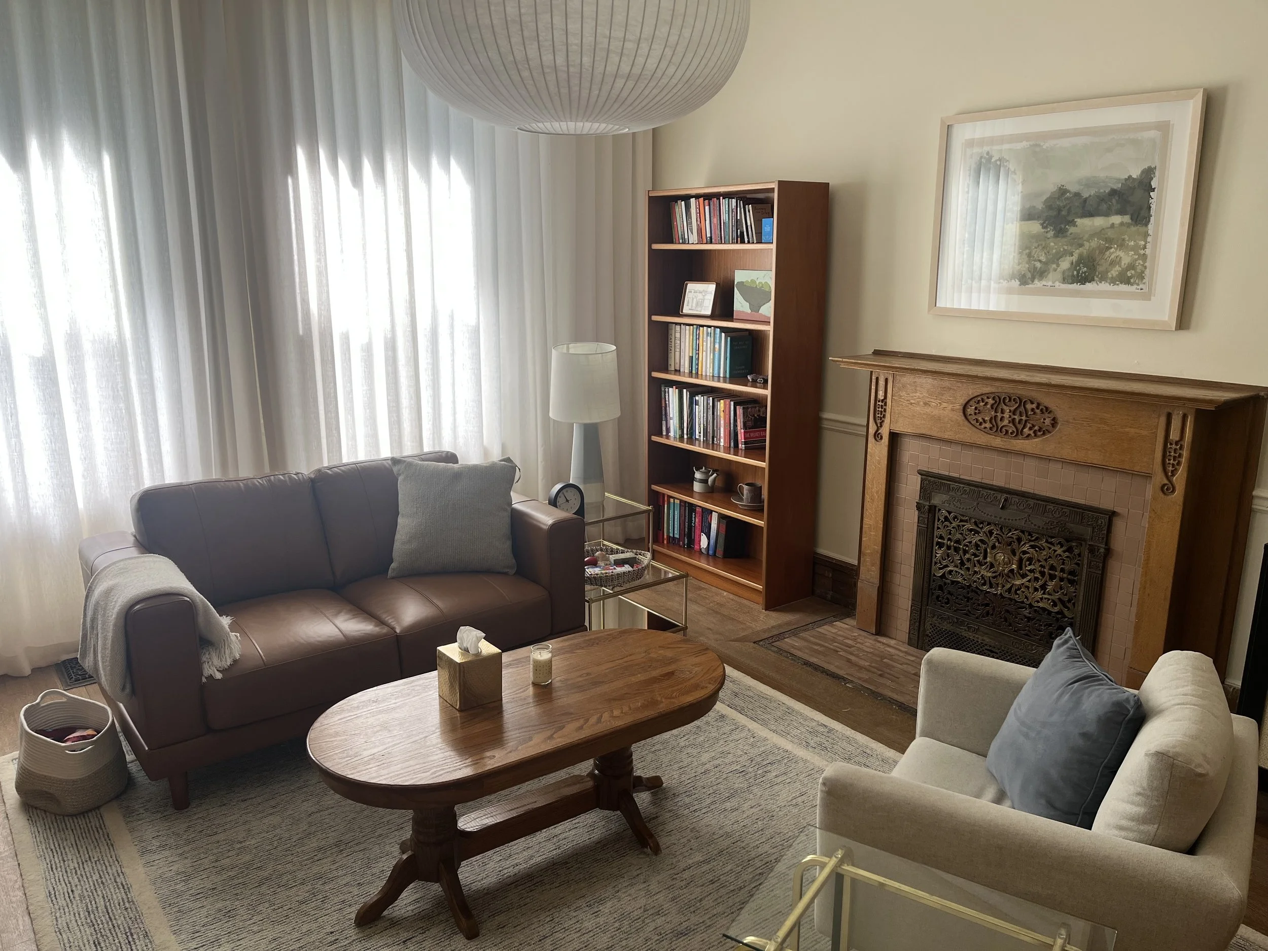 A therapy room with beige walls, a brown leather sofa, a white armchair, a wooden coffee table, tall white curtains, a white floor lamp, a wooden bookshelf, a fireplace with a decorative wooden mantel, and framed artwork on the wall.