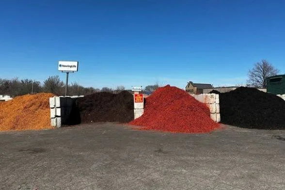 Three piles of colored mulch or soil—orange, dark brown, red, and black—placed next to white concrete blocks on a paved surface with a clear blue sky in the background.