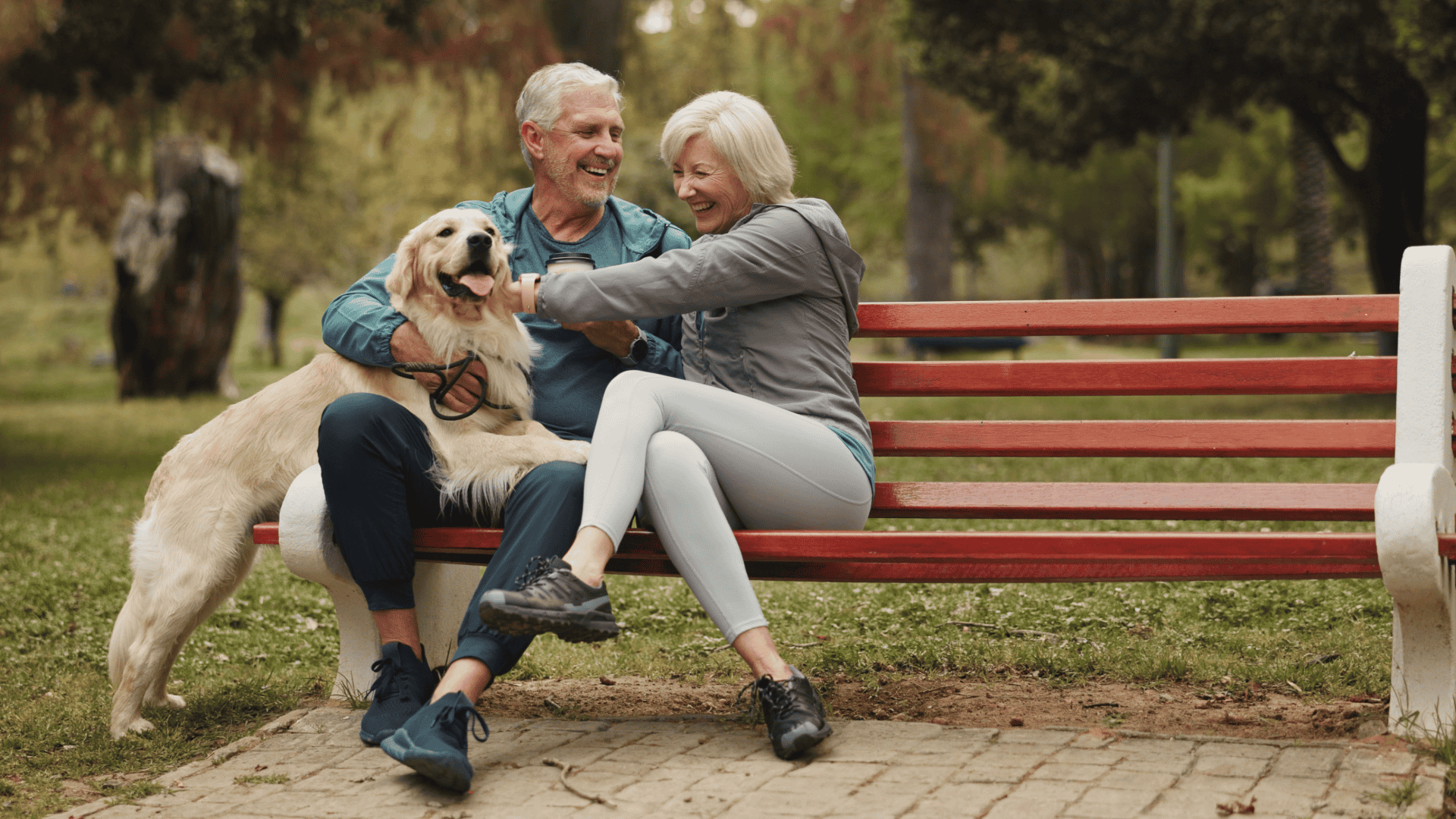An elderly man and woman sitting on a red park bench, smiling and playing with a golden retriever dog in a park with trees in the background.