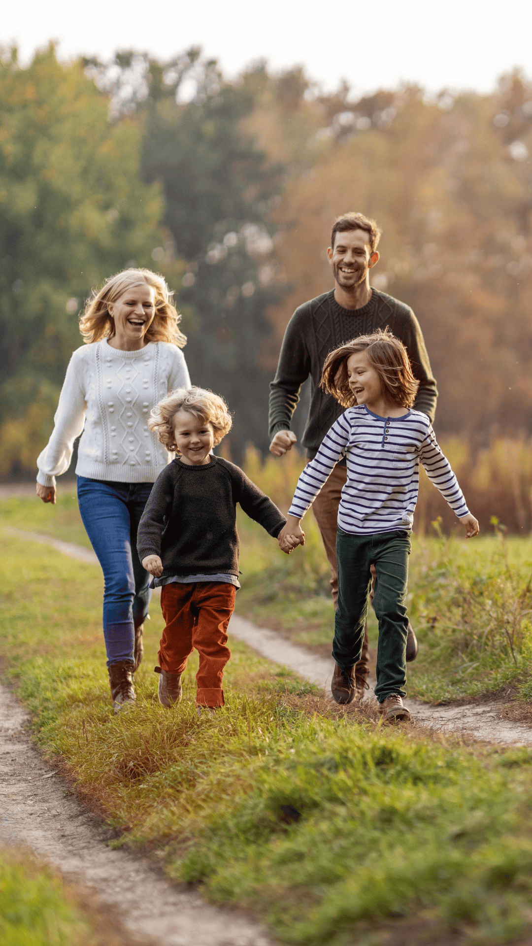 Family of five, including two adults and three children, hiking on a dirt trail through a park with trees in fall foliage, holding hands and smiling.