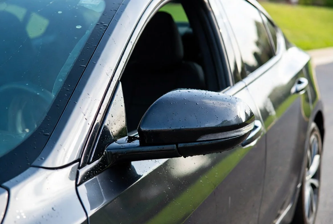 Close-up of a black car's side mirror and front window with raindrops on the surface.
