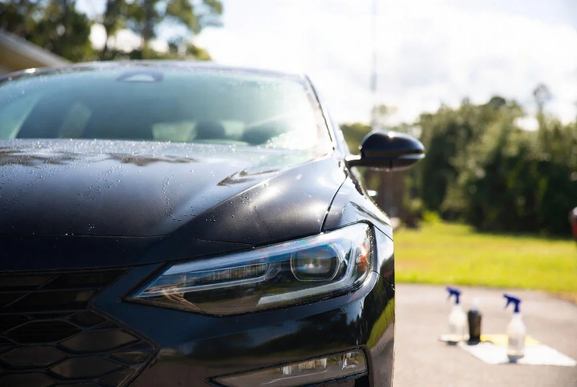 Close-up of a black car with water droplets on its hood, parked outdoors with cleaning supplies in the background.