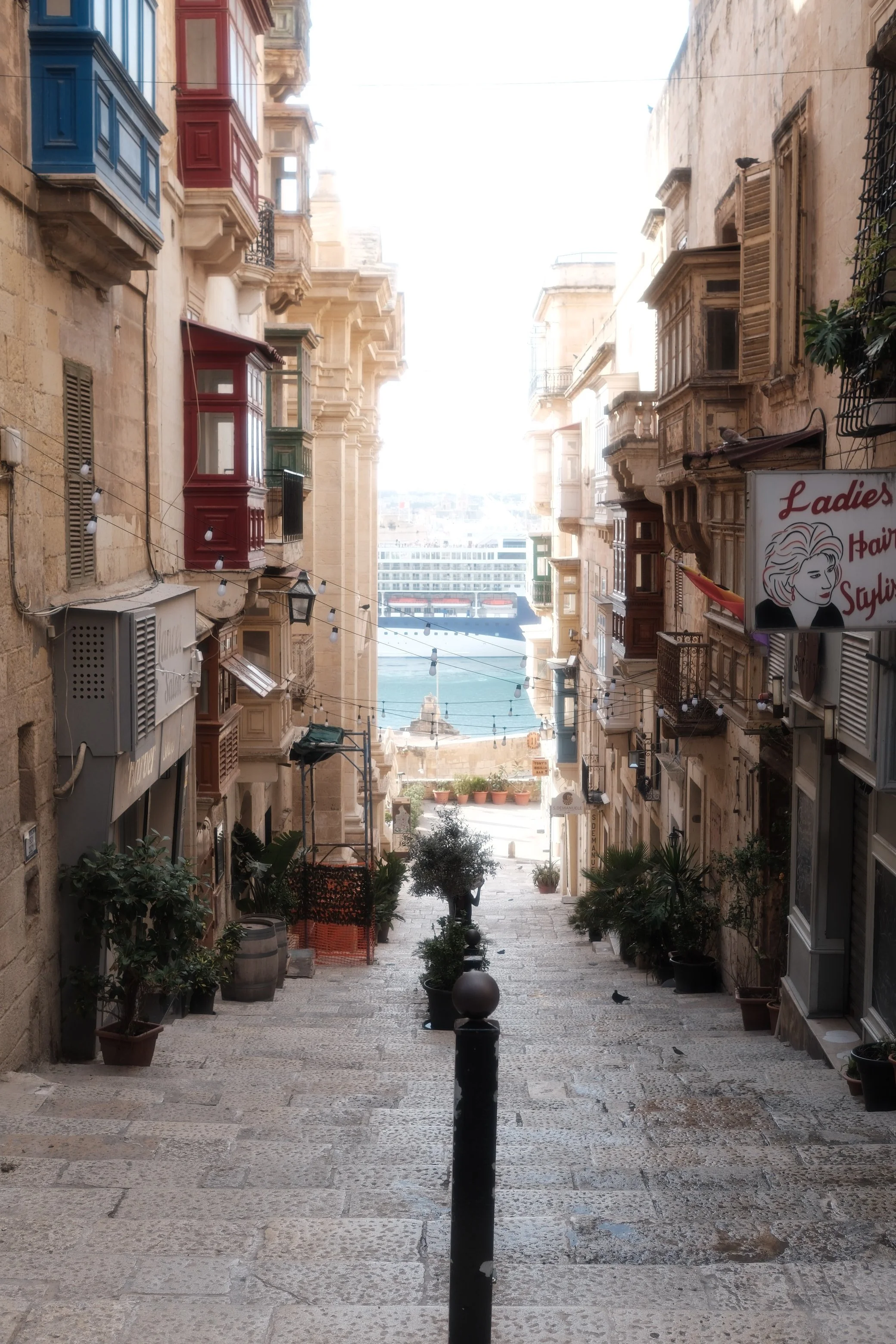 A narrow stone-paved street in a Mediterranean city with decorative balconies and potted plants, leading to a view of water and a large cruise ship in the distance.