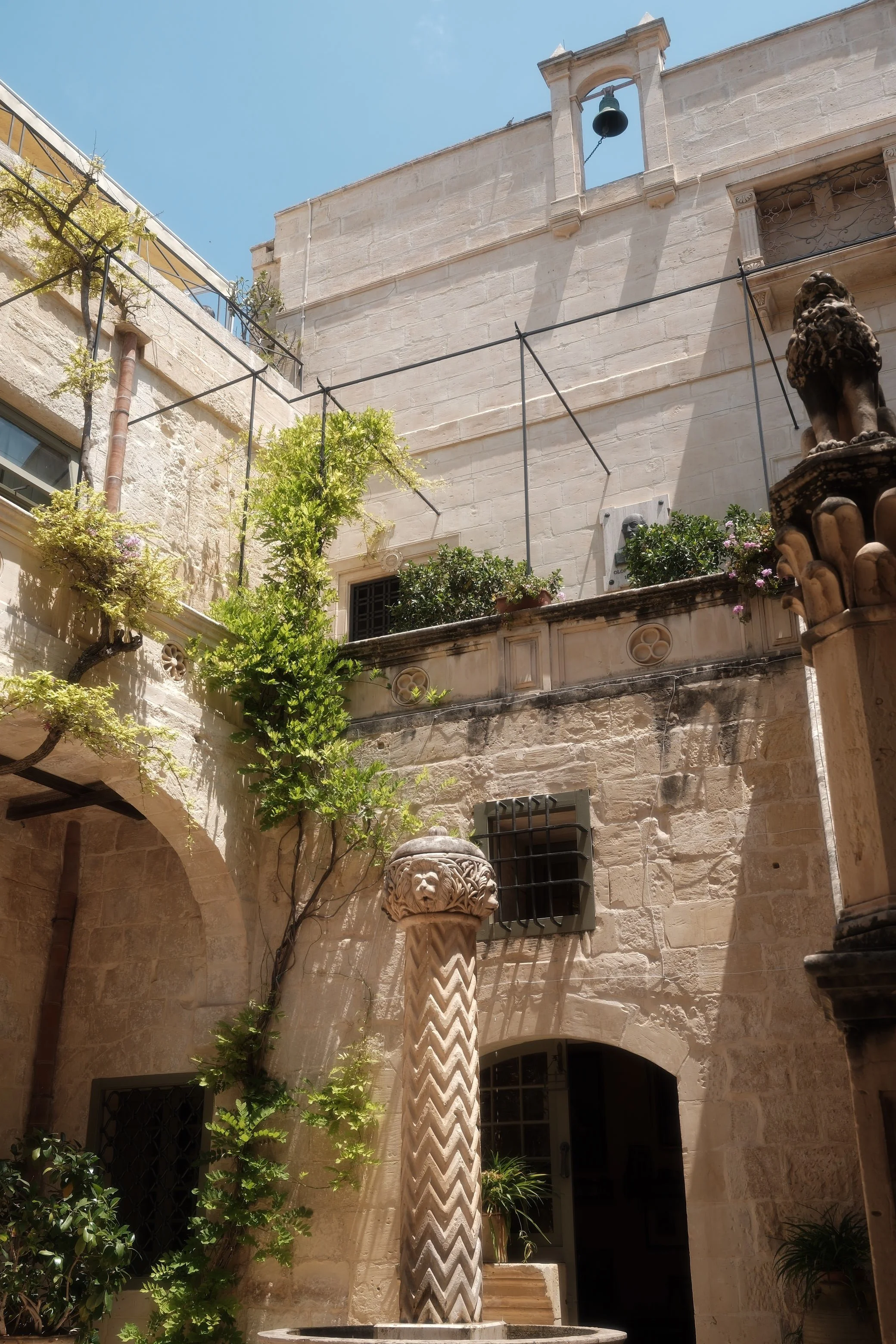 A stone courtyard with a decorative column, climbing plants, potted shrubs, and architectural details including a small bell tower and arched doorway under a clear blue sky.