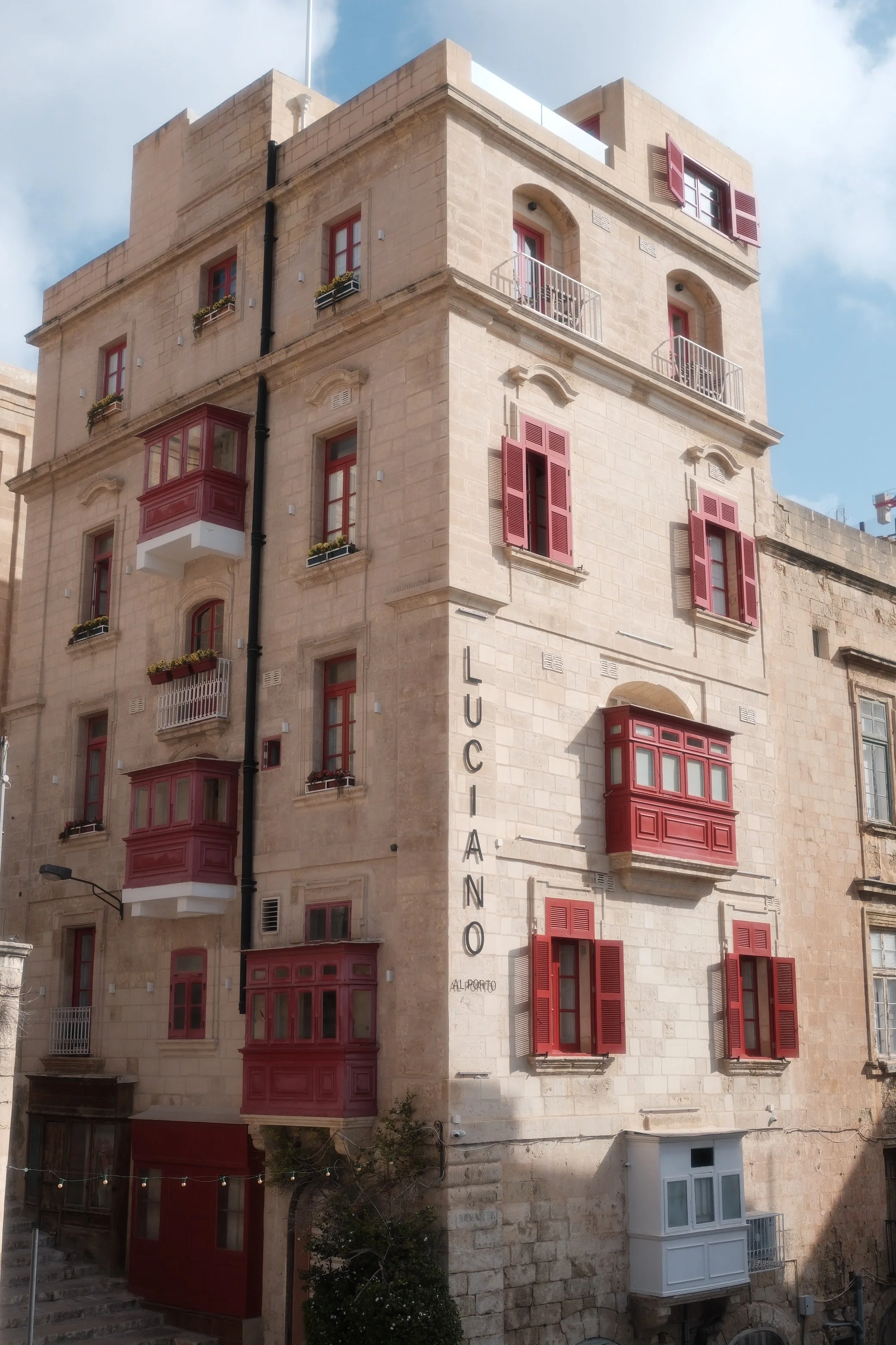 A multi-story beige stone building with red window shutters and balconies, marked with the sign 'LUCIANO' on the side.