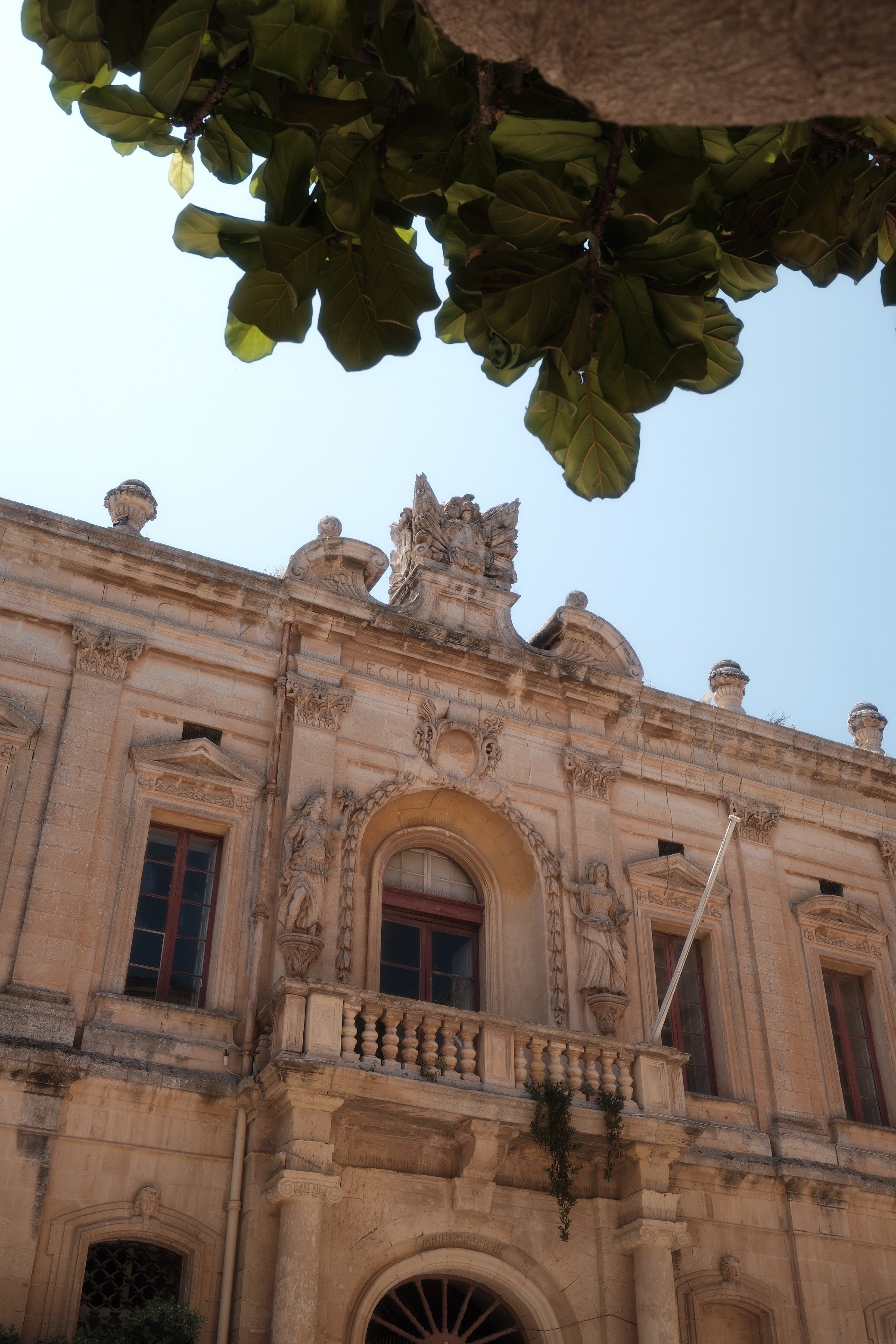 An ornate, historic stone building with decorative columns, statues, and a balcony under a clear blue sky, partially obscured by leafy branches.