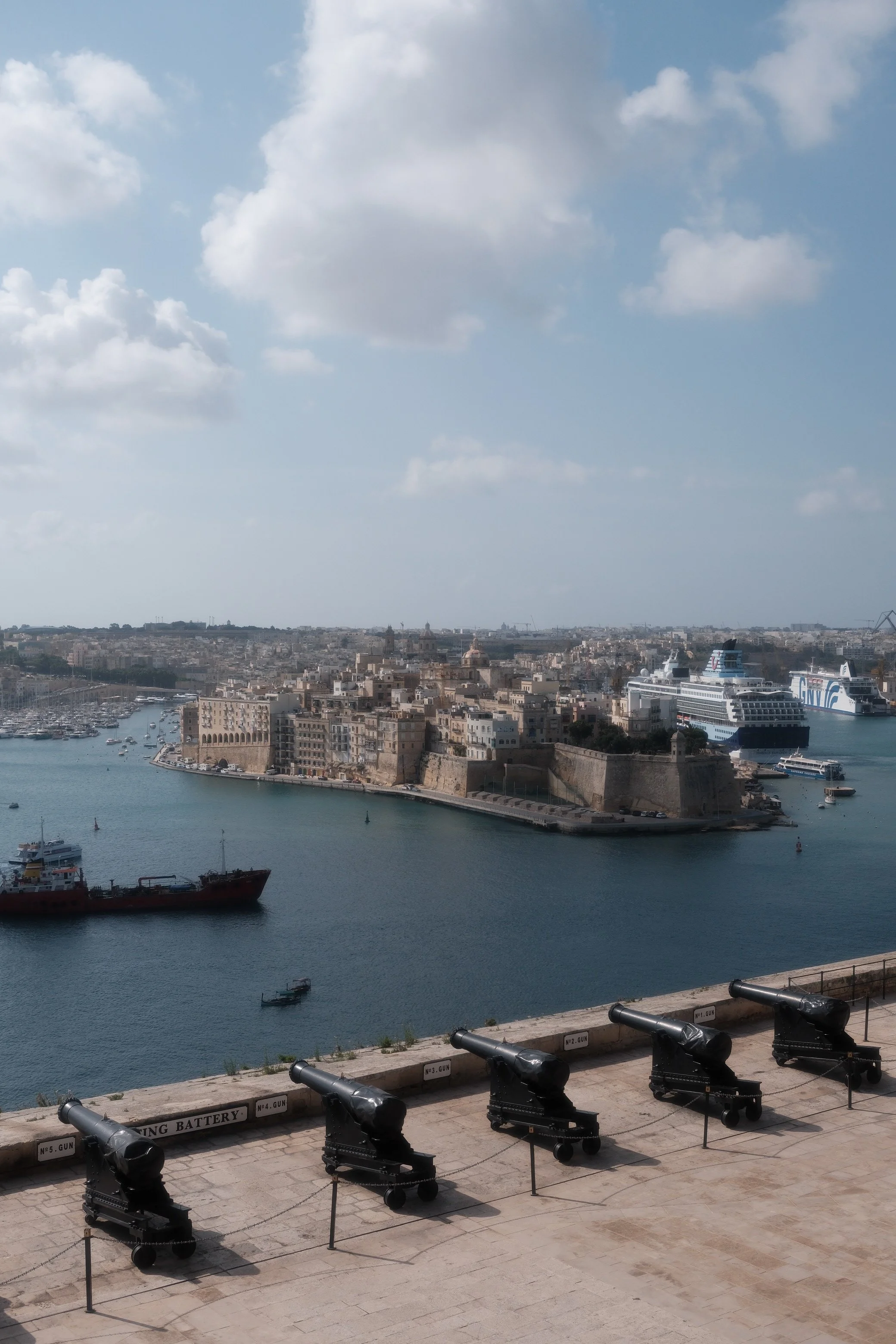 Four cannons facing a harbor with ships and a historic city skyline in the background, under a partly cloudy sky.