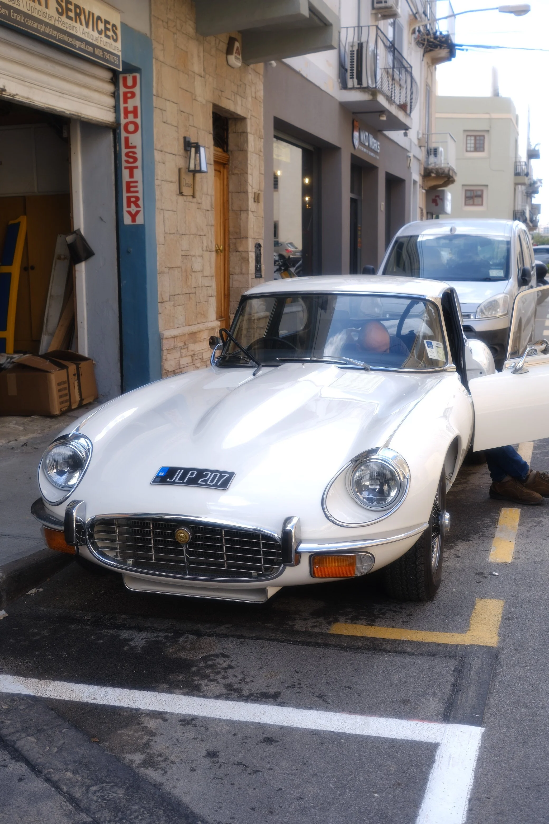 A vintage white sports car parked on the street, with a black license plate reading 'JLP 207', nearby a modern silver SUV, in front of a building with a sign for upholstery and repair services.
