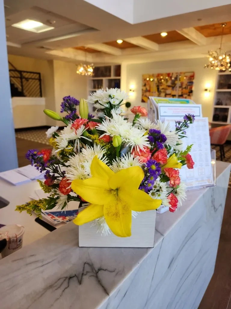 Colorful flower arrangement with yellow lily, white daisies, pink, purple, and green flowers on a marble counter in a well-lit lobby or reception area.