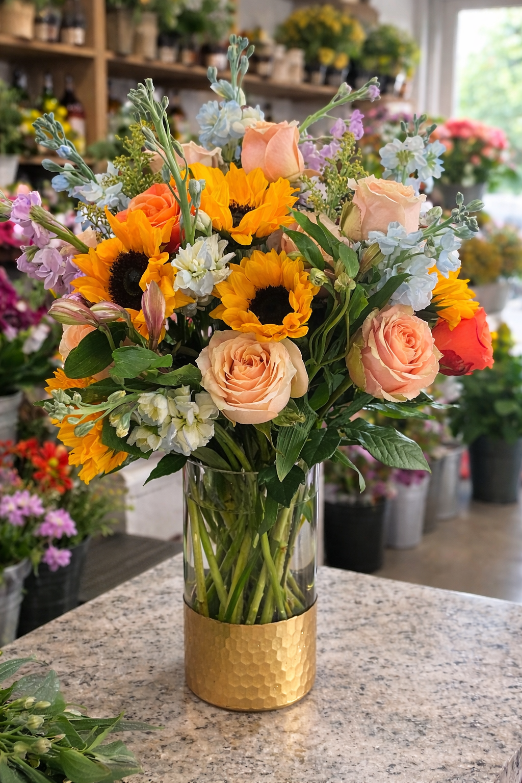 A colorful bouquet of sunflowers, peach roses, and white, blue, and pink flowers in a clear glass vase with a gold-colored band, St. George UT delivery.