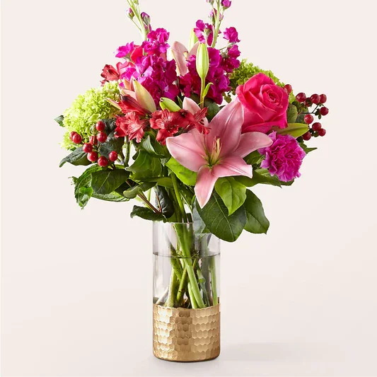 Colorful mixed flower bouquet in a clear glass vase with gold accent, containing pink roses, lilies, and other vibrant flowers.