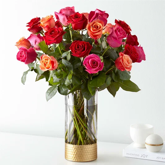 A clear glass vase with a gold base filled with a large bouquet of pink, red, and orange roses, placed on a white surface with white cups and a tray nearby.