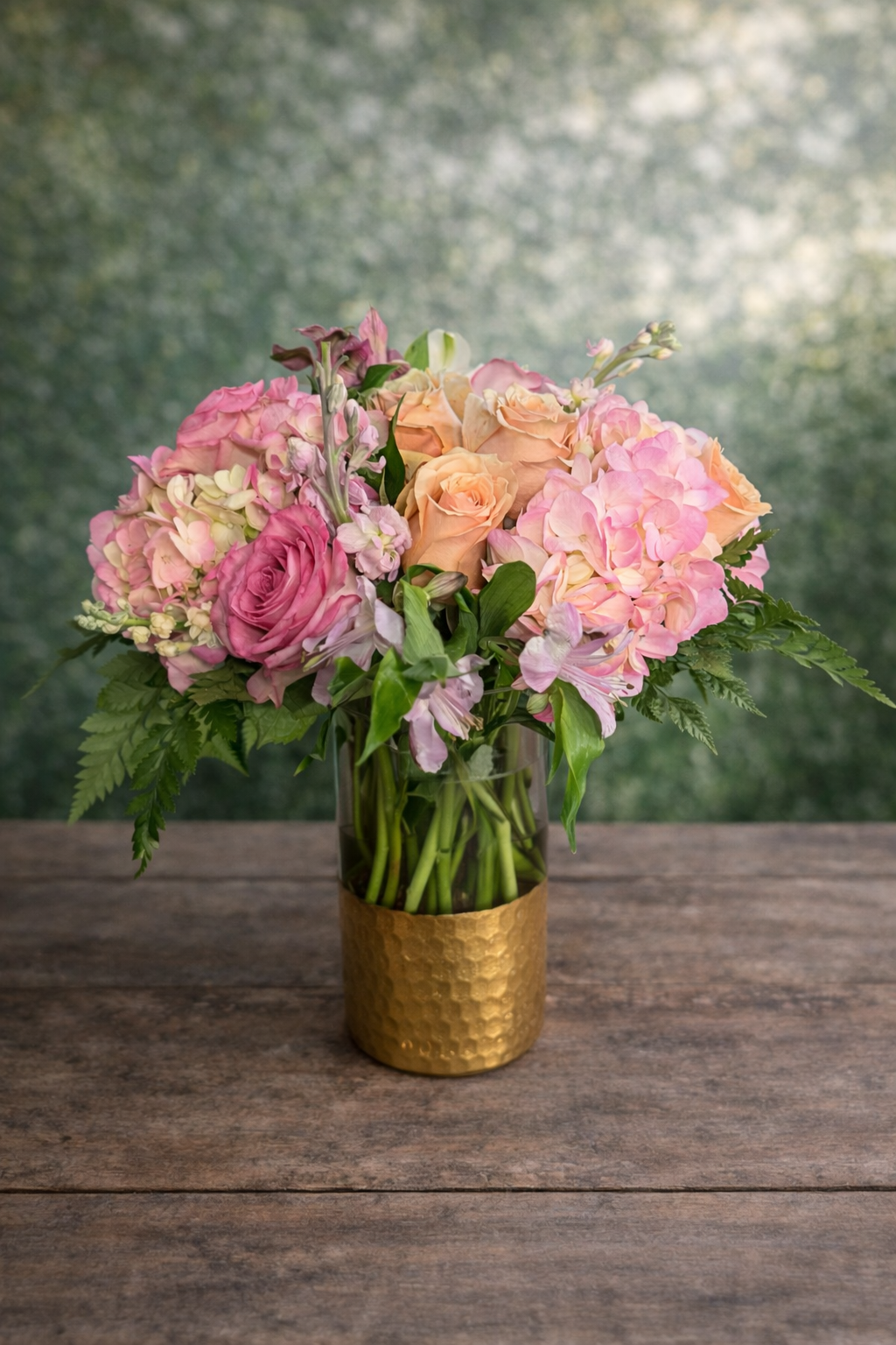 A floral arrangement of pink roses, hydrangeas, and other flowers in a clear glass vase with a gold textured base, St. George Delivery