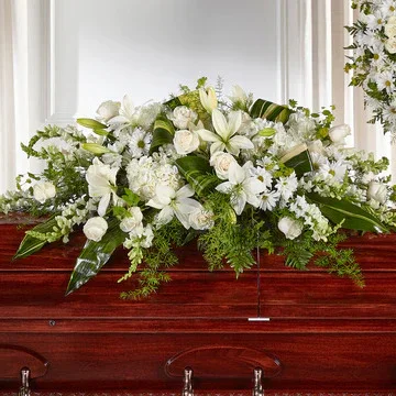 A white floral casket spray with lilies, roses, and greenery resting on a wooden casket.
