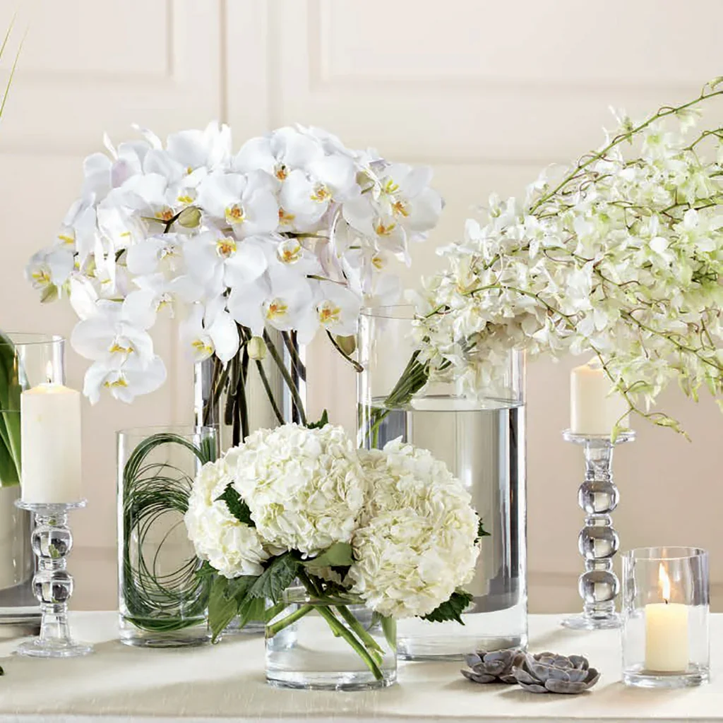 Arrangement of white orchids, hydrangeas, and other white flowers in glass vases with candles on a table.