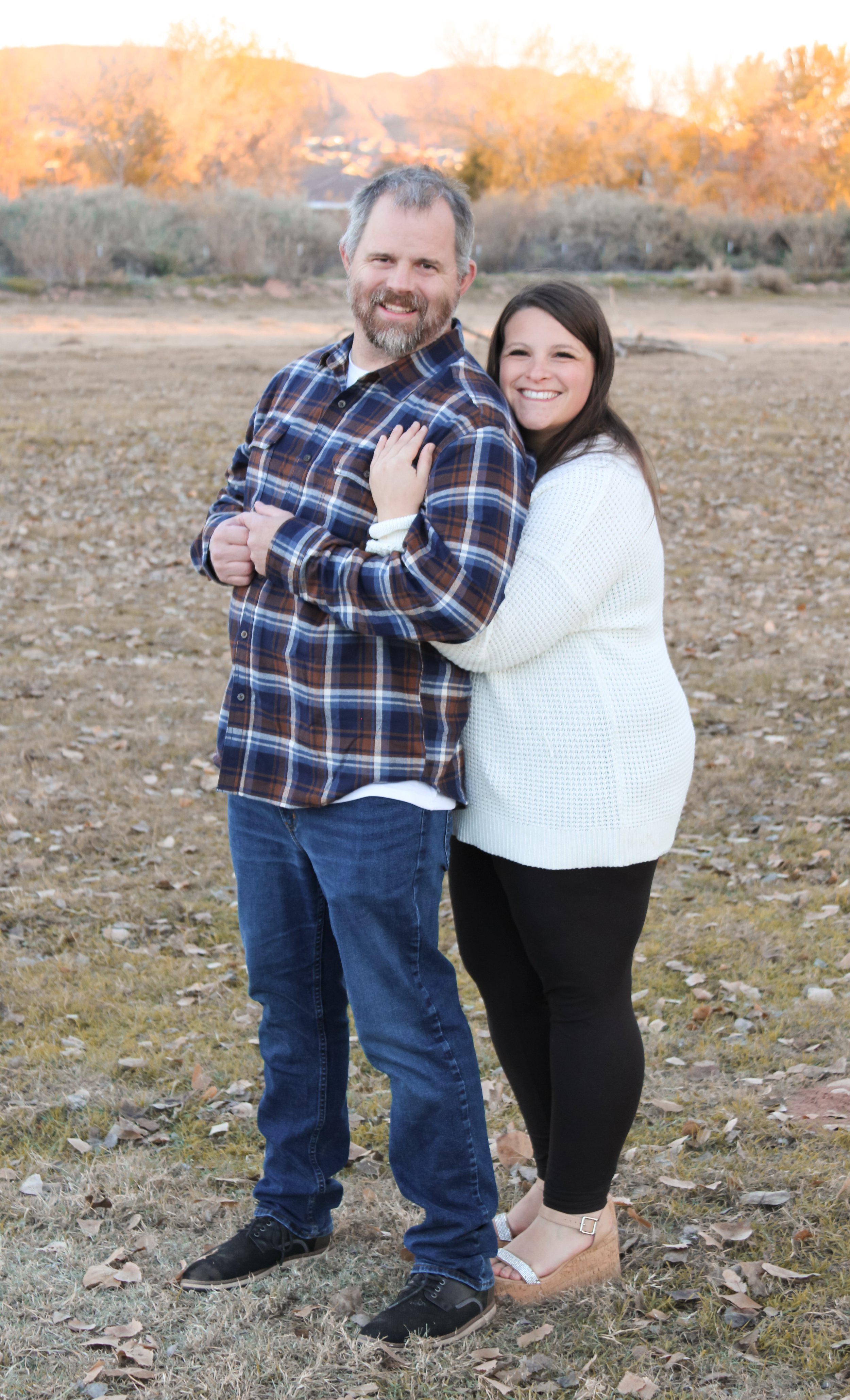 A man and a woman standing outside on a fall day, smiling and embracing, with trees in autumn colors in the background.