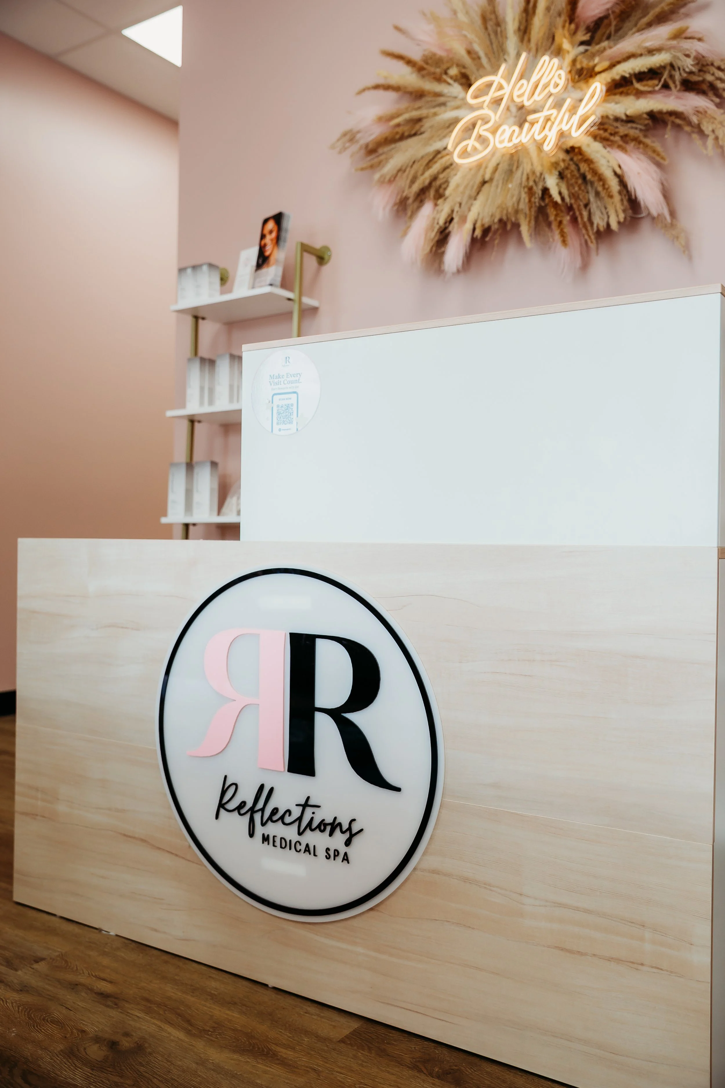 Reception area of a medical spa called 'Reflections Medical Spa' with a sign on the front desk featuring the spa's logo and name, a pink neon sign above that reads 'Hello Beautiful', and decorative dried plants on a wall.
