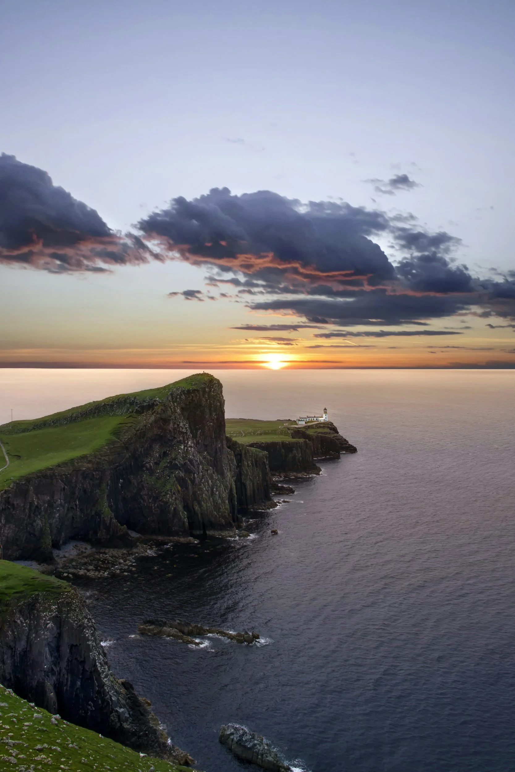 A coastal scene at sunrise with a lighthouse on a cliff, surrounded by green grassy areas, dark clouds in the sky, and calm ocean waters.