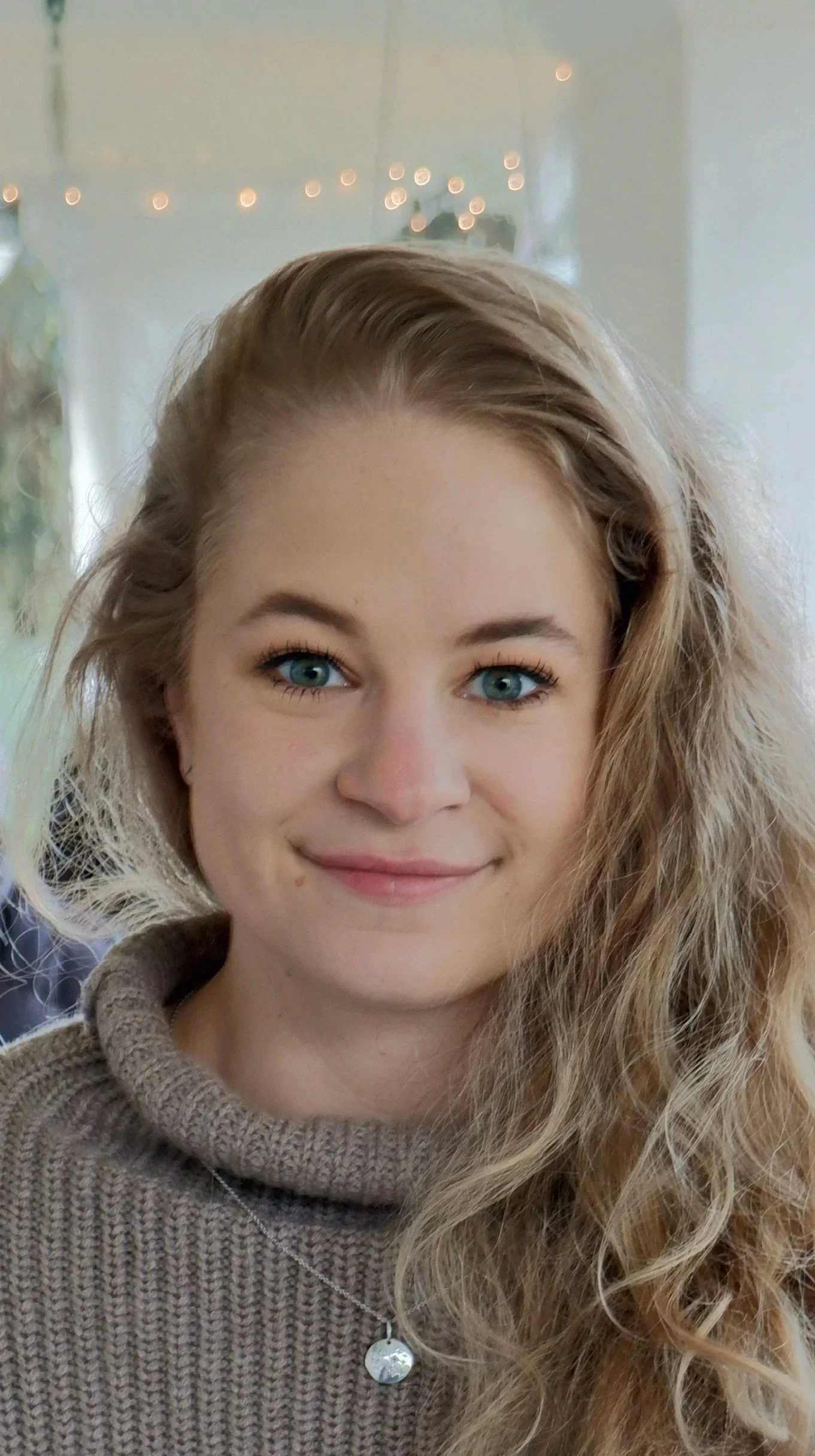 Close-up of a young woman with blue eyes, long curly blonde hair, wearing a gray knit sweater and a silver necklace with a round pendant, smiling in a well-lit room.