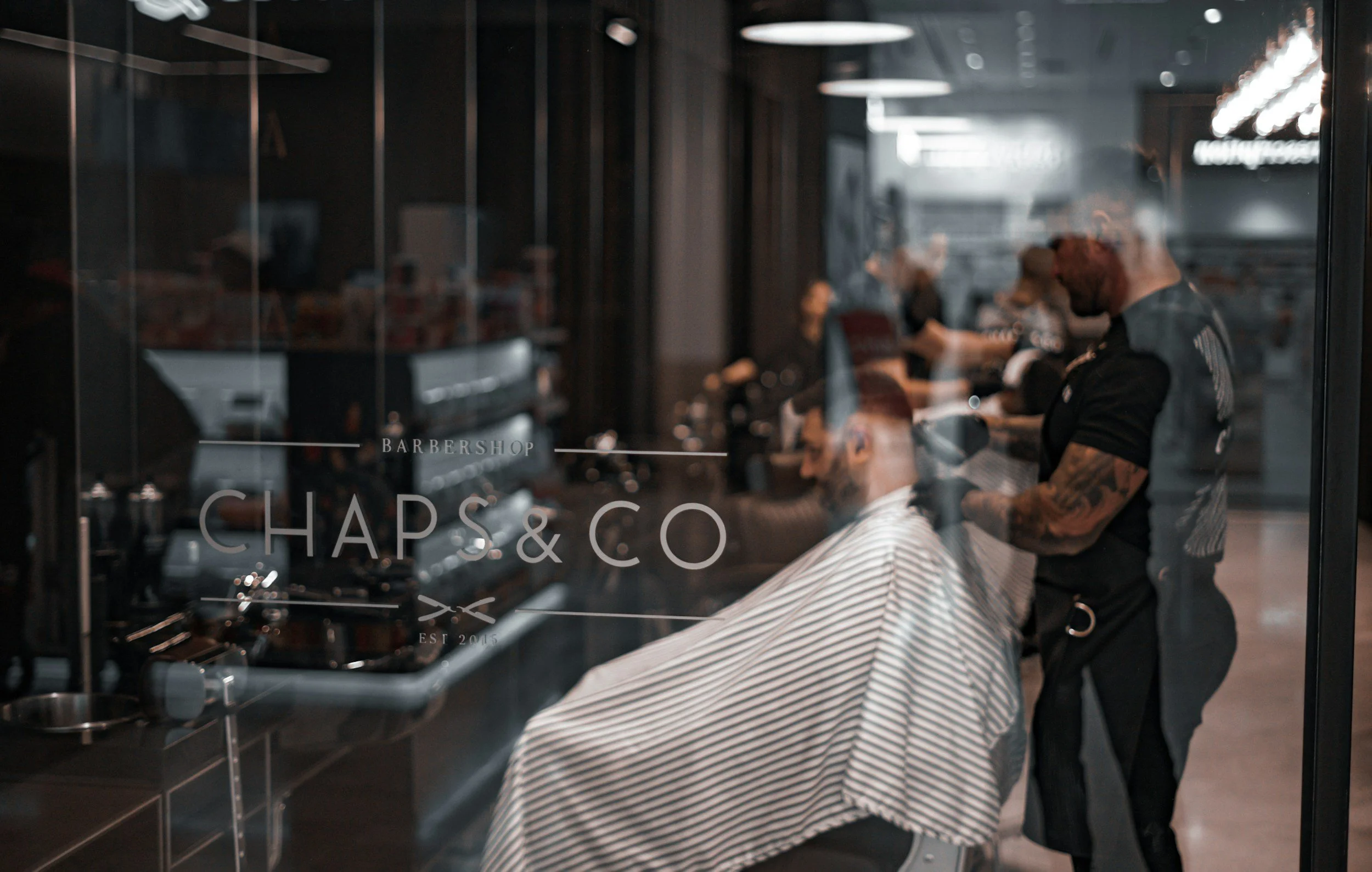Inside a barbershop with a barber cutting a man's hair, who is seated and covered with a striped cape. The shop has glass walls with the