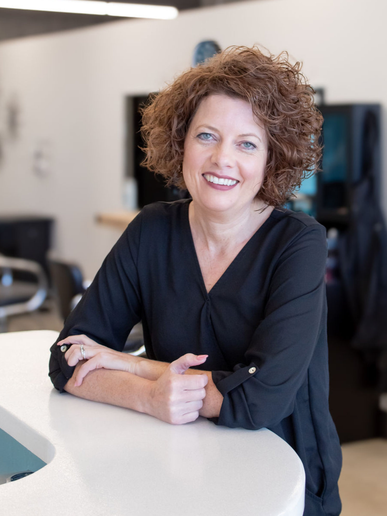 Woman with curly red hair wearing a black blouse, smiling and leaning on a white reception desk in an office setting.
