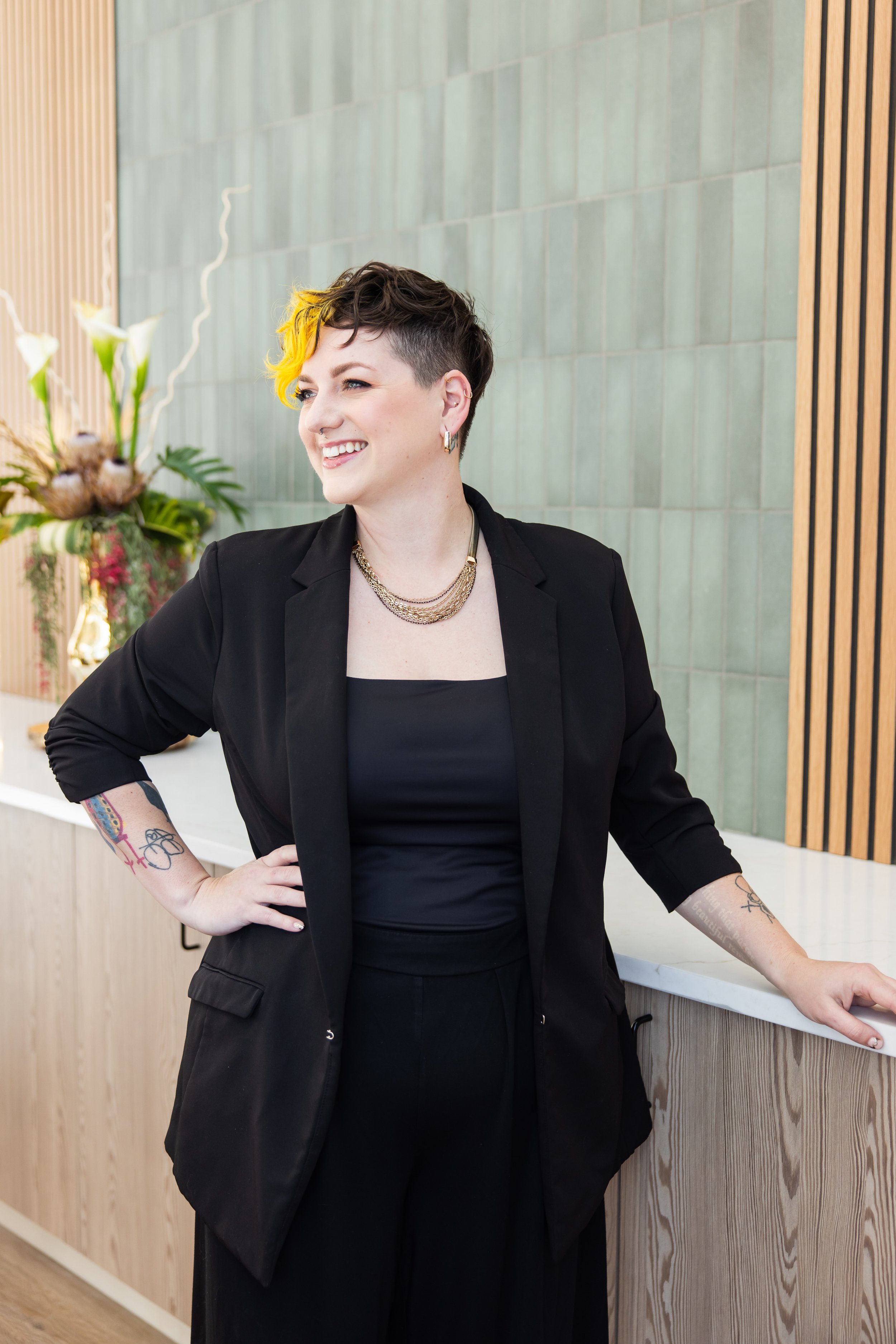 A woman with short, dark hair and yellow highlights, wearing a black blazer and jewelry, smiling and standing at a reception desk.