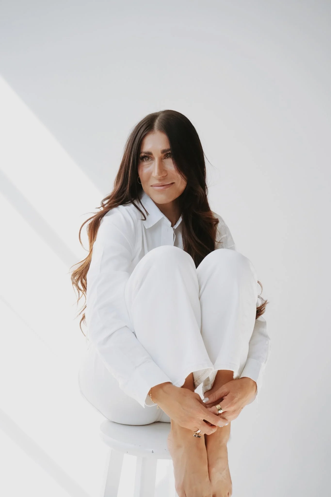 Woman with long dark hair, sitting on a white stool, wearing an all-white outfit, with her knees drawn up and hands holding her feet, against a bright white background.