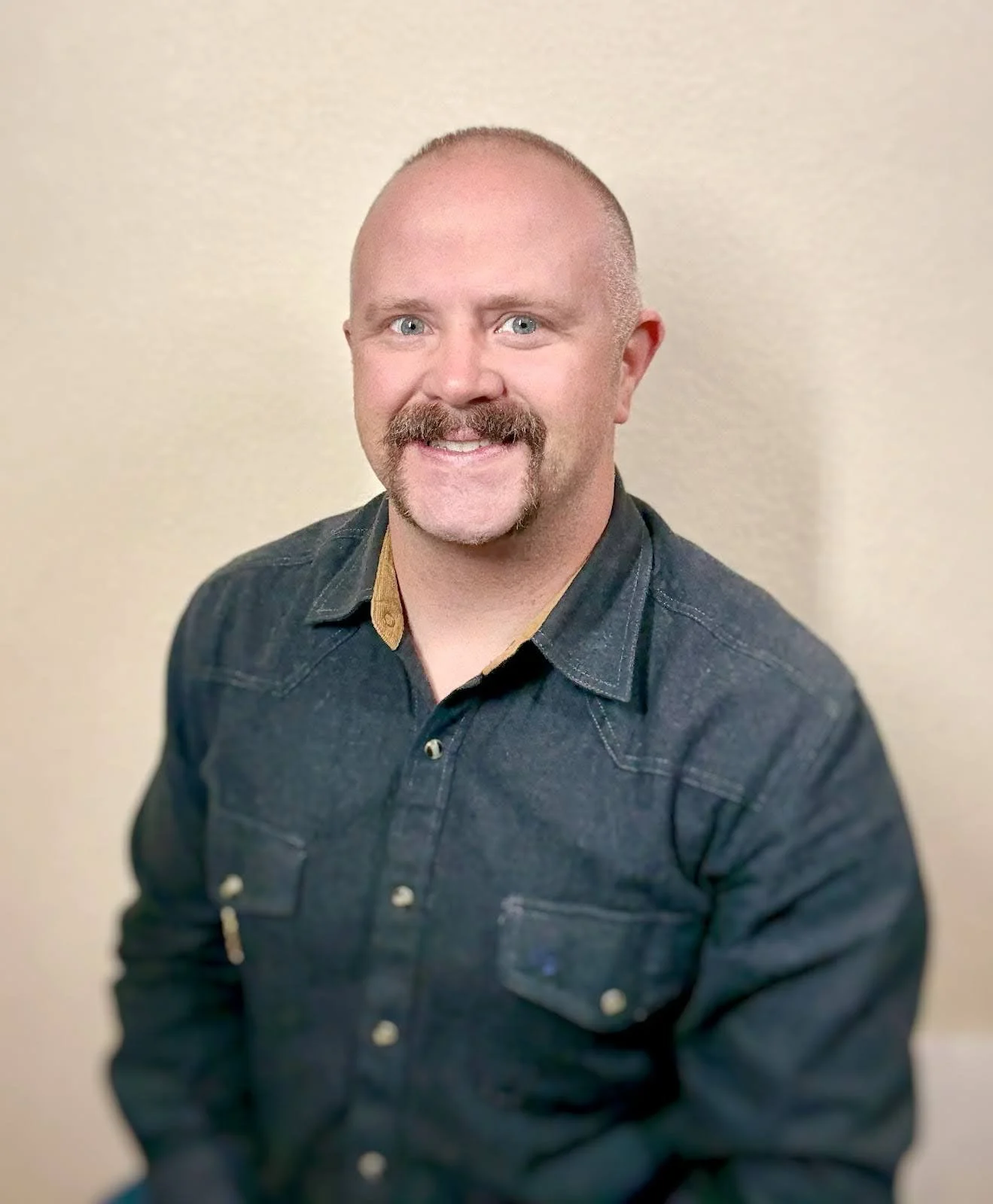 A man with a shaved head, blue eyes, and a goatee, smiling, wearing a black denim shirt with a tan collar, against a plain beige background.