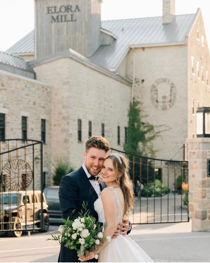 Bride and groom smiling in front of Elora Mill with bouquet, wedding attire, and stone building background.