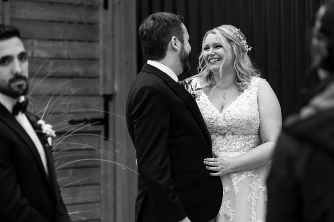 Bride and groom smiling during outdoor wedding ceremony with groomsman in background.