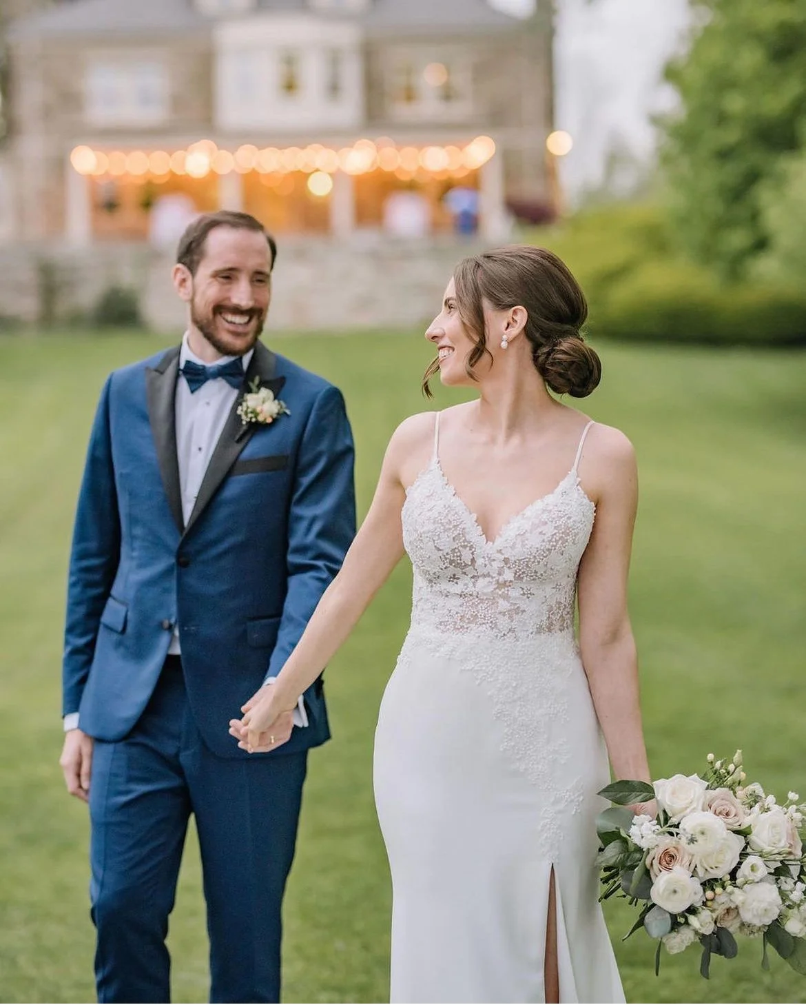 Bride and groom holding hands outdoors. The bride wears a lace wedding dress and holds a bouquet of flowers. The groom is in a blue suit with a bow tie. A lit building is in the background.