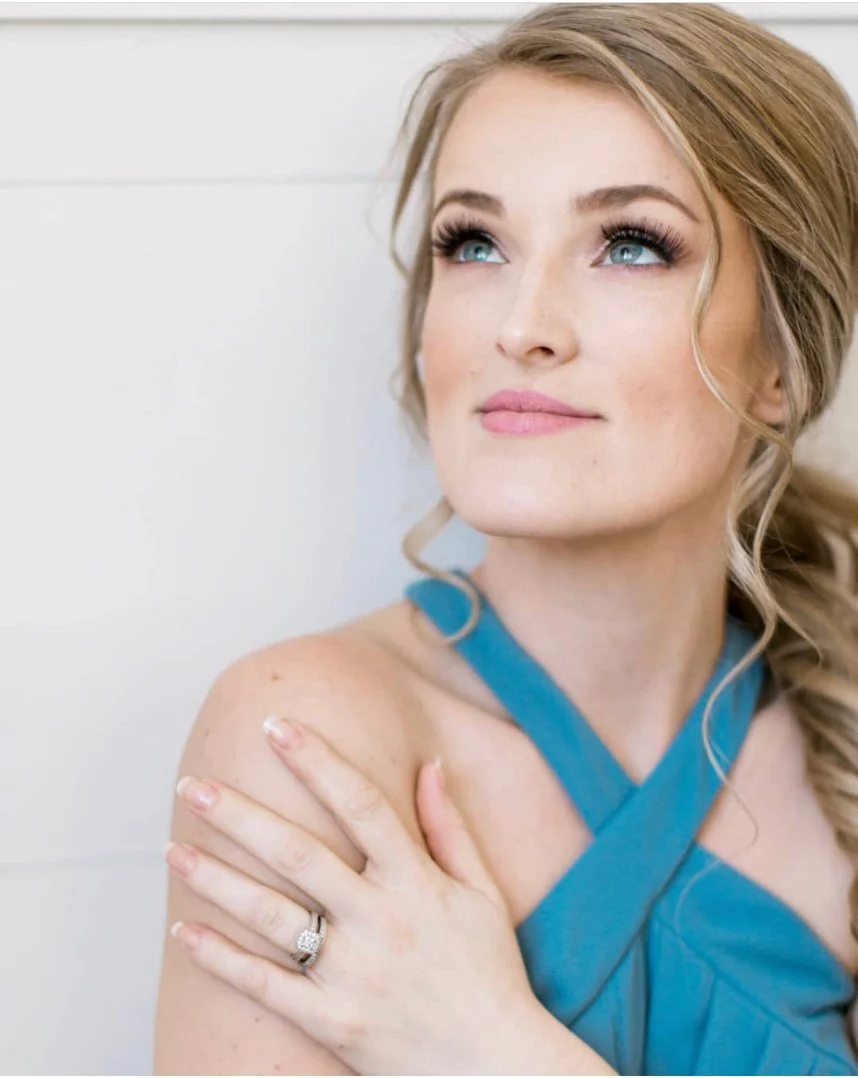 Woman in a blue dress with crossed straps, looking upwards with a gentle expression, wearing a ring on her left hand.