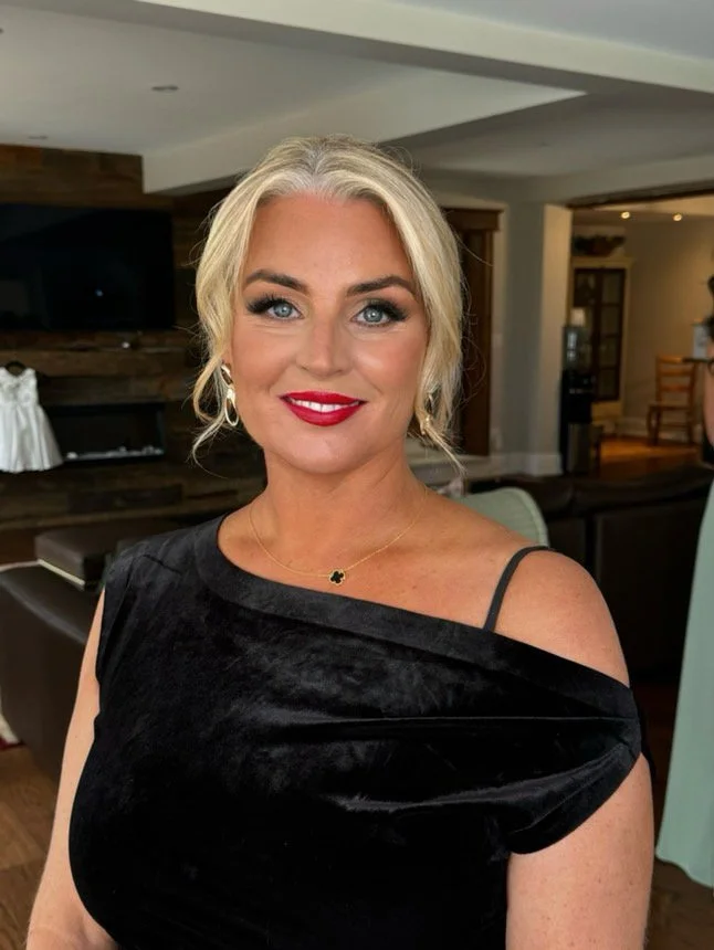 Woman with blonde hair and red lipstick wearing a black dress, standing in a living room.