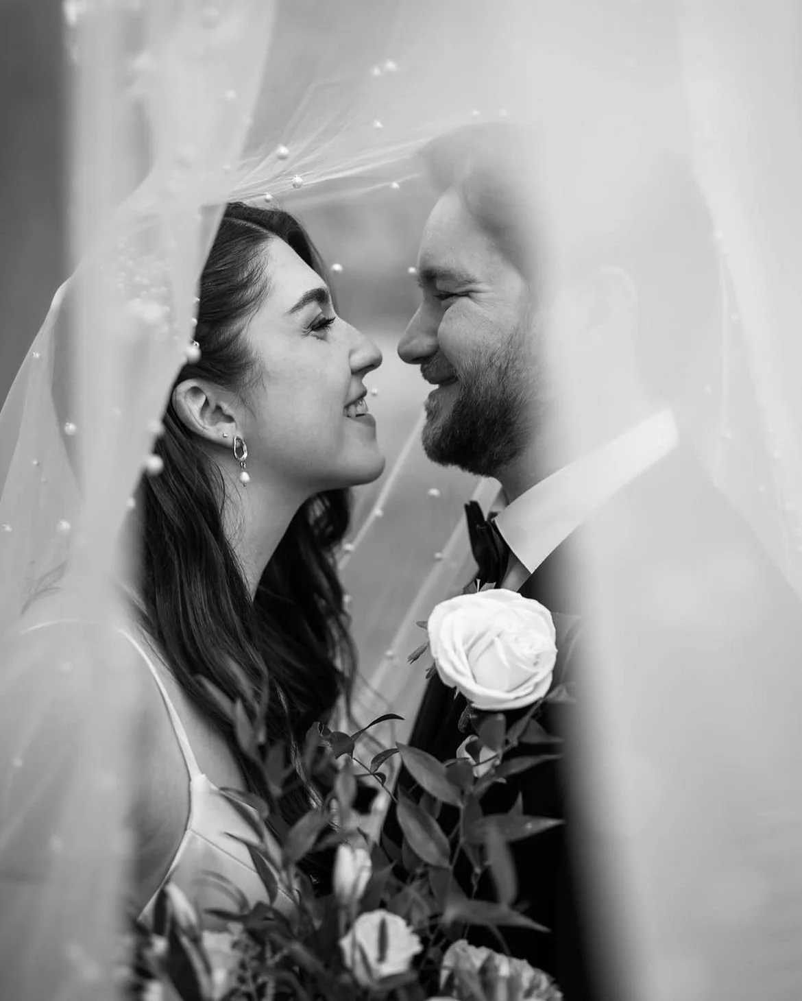 Romantic black and white photo of a wedding couple under a veil, smiling at each other, holding flowers.