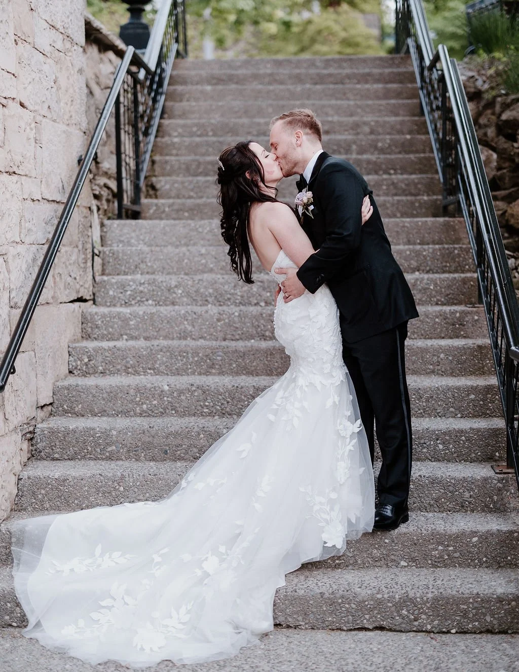 Bride and groom kissing on stone staircase, with the bride in a white dress and groom in a black suit.
