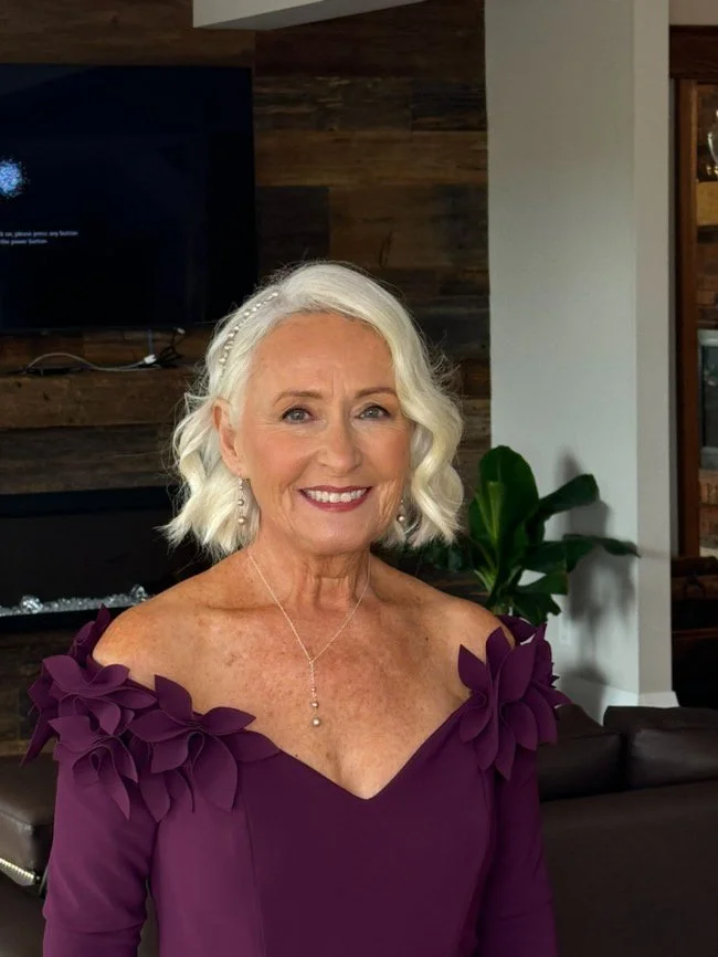 Older woman with white hair wearing a purple off-shoulder dress inside a room.