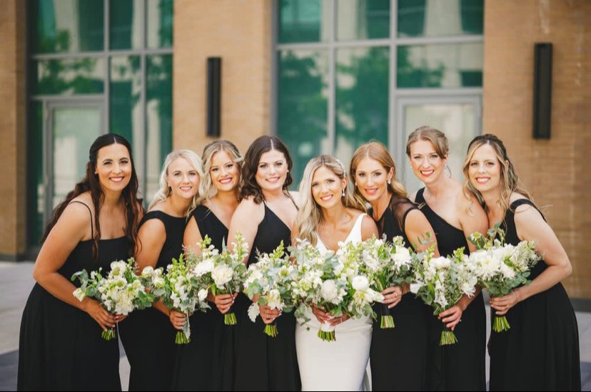 A bride and seven bridesmaids in black dresses holding white and green floral bouquets, standing in front of a building.