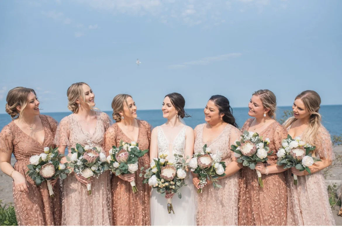 Bride and bridesmaids holding bouquets, wearing matching dresses, with a beach background.