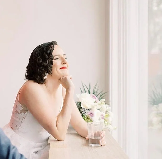 Smiling woman in white dress with short dark hair holding a glass, next to a bouquet of flowers on a wooden surface.