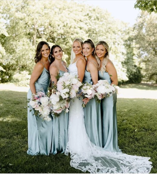 A bride in a white gown with a lace train poses with four bridesmaids in matching light blue dresses, each holding bouquets of flowers, in an outdoor grassy area with trees in the background.