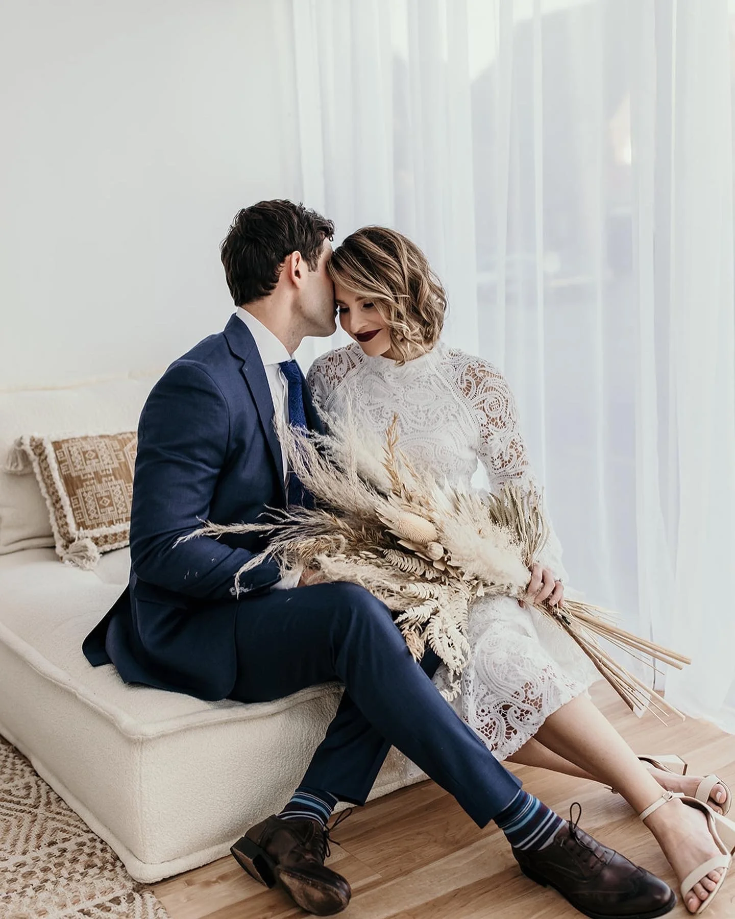 A couple sitting on a couch; the man is wearing a blue suit and the woman is in a white lace dress with a bouquet of dried flowers.