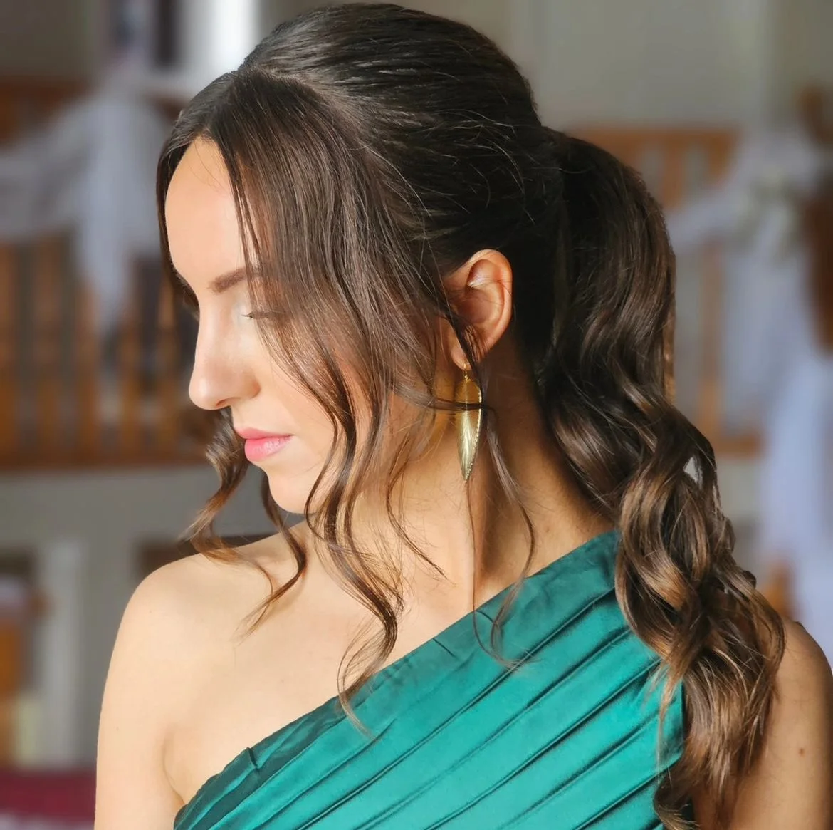 Woman with long brown hair in an elegant ponytail, wearing an off-the-shoulder emerald green gown and gold earrings, looking down in a softly lit room.