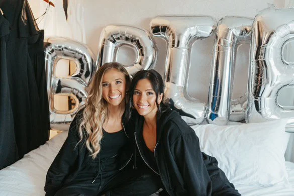 Two women sitting on a bed with "BRIDE" silver balloons in the background.