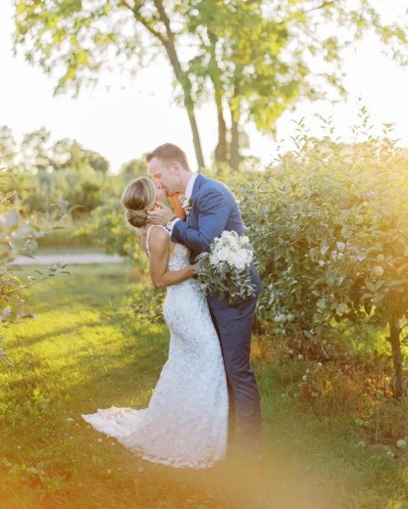 Bride and groom kissing in a sunlit garden, holding a bridal bouquet.