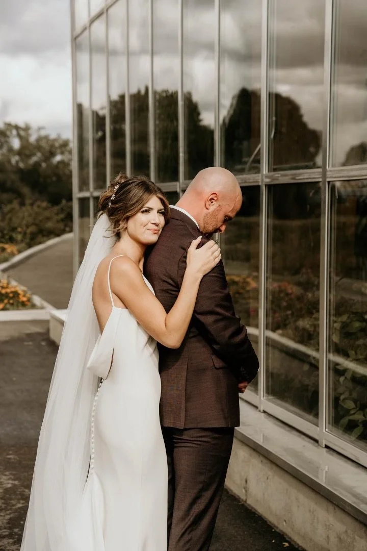 Bride hugging groom from behind near glass building.