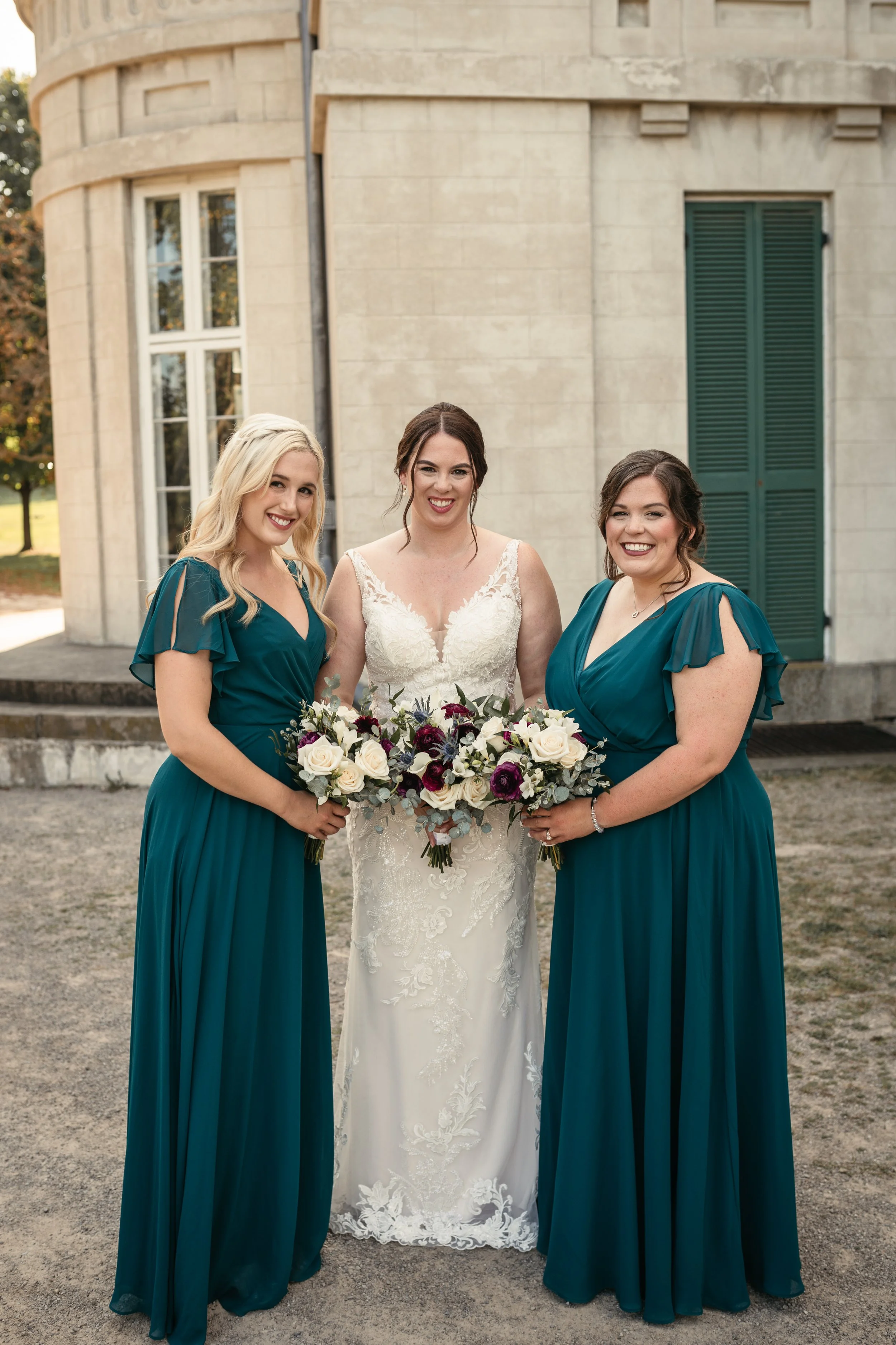 Bride with two bridesmaids in teal dresses, holding bouquets, standing in front of a stone building.