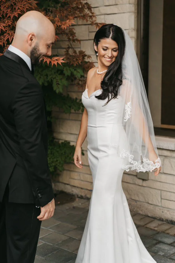 Bride in white wedding dress with veil, smiling near groom in black suit, standing outside with brick wall and foliage background.