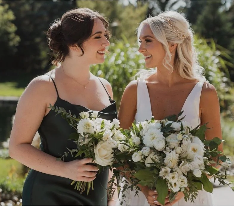Two women holding bouquets, smiling at each other, one in a white dress and the other in a dark dress, outdoors.