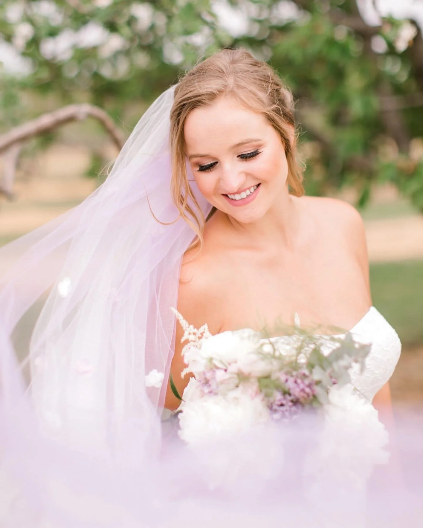 Bride in strapless dress holding a bouquet, smiling under veil outdoors.